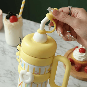 Yellow children's insulated thermos flask cup with a handle and straw attachment, held by a hand, with desserts in the background.