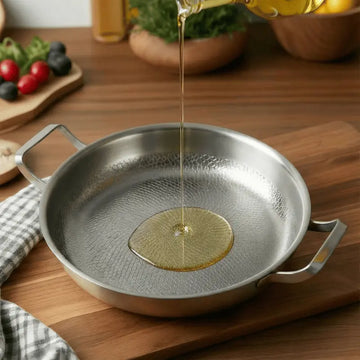 Oil being poured into a Titanium Hammered Double-Handle Frying Pan on a wooden surface with vegetables in the background