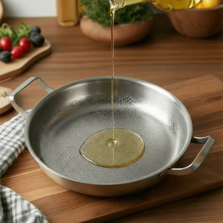 Oil being poured into a Titanium Hammered Double-Handle Frying Pan on a wooden surface with vegetables in the background